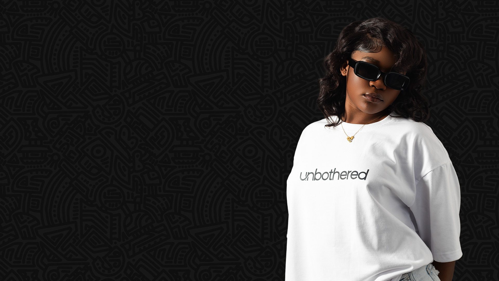 Model wearing the white Unbothered oversized tee and black sunglasses against an African line-pattern backdrop